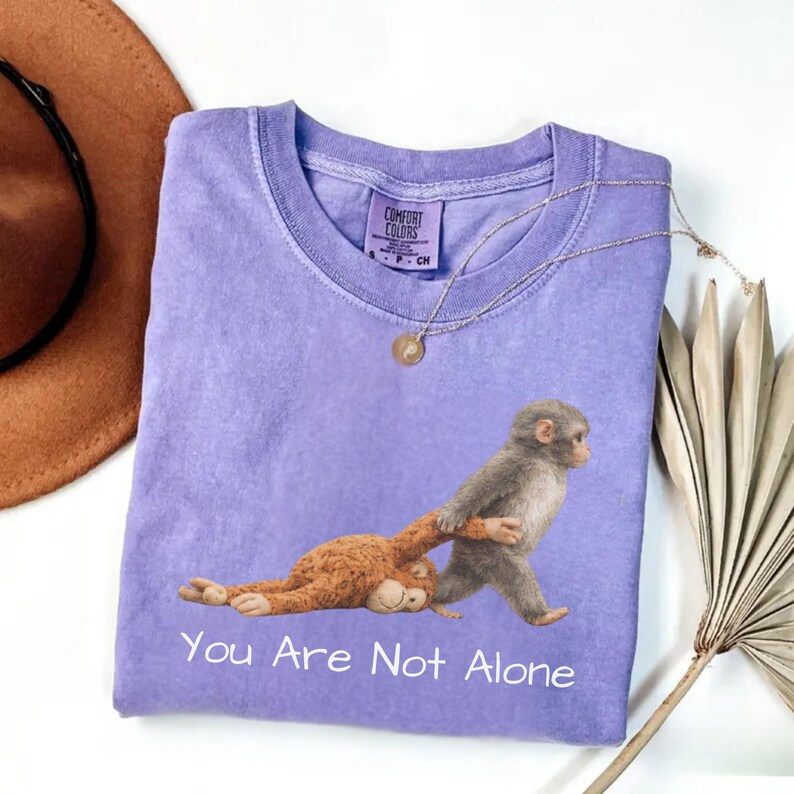 Puede incluir: Una camiseta lila Comfort Colors con un gr&aacute;fico de dos monos, uno tirando del otro. El texto "You Are Not Alone" est&aacute; impreso debajo. Un collar dorado y un sombrero marr&oacute;n son visibles.