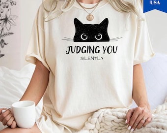 May include: Ivory-colored t-shirt featuring a black cat graphic with the text "JUDGING YOU SILENTLY." The cat's eyes are visible above the text. The shirt is a relaxed fit, with rolled sleeves. The shirt is made of a soft material.