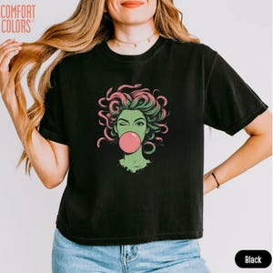 May include: Black t-shirt with a graphic of Medusa winking and blowing a bubblegum bubble. The Medusa illustration is green and pink. The shirt has the words "Comfort Colors" printed on the upper left.