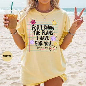 May include: A butter yellow short-sleeve t-shirt featuring a graphic design of lined notebook paper with the text "FOR I KNOW THE PLANS I HAVE FOR YOU" and "Jeremiah 29:11". The design includes colorful flowers, stars, and a smiling face icon.