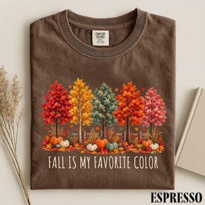 May include: A brown short-sleeve t-shirt featuring a graphic of five autumn trees in shades of red, orange, yellow, and green, with pumpkins at the base. The text "FALL IS MY FAVORITE COLOR" is printed below the trees. A Comfort Colors label is visible.