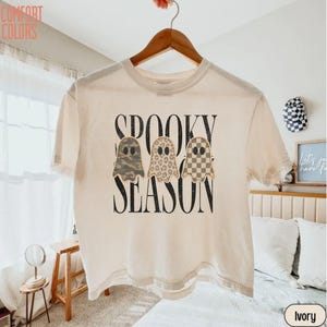 May include: An ivory short-sleeve t-shirt featuring the black text SPOOKY SEASON and three ghost illustrations. The ghosts have distinct patterns: one in camouflage, one in leopard print, and one in a tan and white checkered pattern.