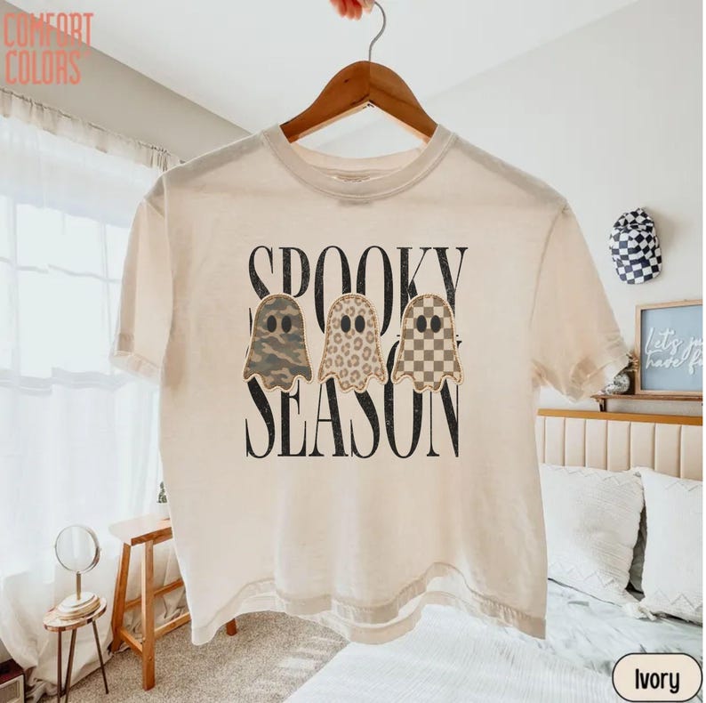 May include: An ivory short-sleeve t-shirt featuring the black text SPOOKY SEASON and three ghost illustrations. The ghosts have distinct patterns: one in camouflage, one in leopard print, and one in a tan and white checkered pattern.