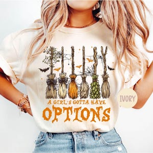 May include: An ivory t-shirt featuring a graphic of six different witch brooms arranged to spell the word WITCH. The design includes spiderwebs, bats, and the orange text A GIRL'S GOTTA HAVE OPTIONS. The shirt has short sleeves and a crew neckline.
