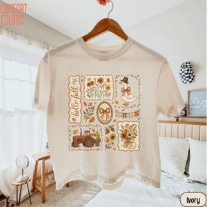 May include: An ivory short-sleeve t-shirt featuring a graphic print with the text "hello fall" and various autumn-themed illustrations including a tractor, a goose wearing a hat, sunflowers, pumpkins, and decorative floral borders in warm tones.