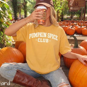 May include: A mustard yellow short-sleeved t-shirt featuring the white text PUMPKIN SPICE CLUB printed on the chest. The shirt is worn with light blue distressed denim jeans and brown boots, while a brown baseball cap is also visible.