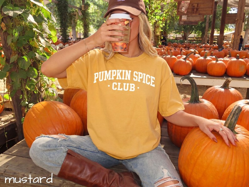 May include: A mustard yellow short-sleeved t-shirt featuring the white text PUMPKIN SPICE CLUB printed on the chest. The shirt is worn with light blue distressed denim jeans and brown boots, while a brown baseball cap is also visible.