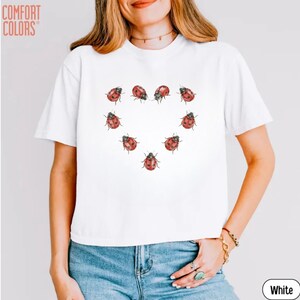 May include: A white short-sleeved t-shirt featuring a graphic print of red and black ladybugs arranged in the shape of a heart on the center chest. The text COMFORT COLORS is printed in the upper left corner. The shirt has a crew neckline.