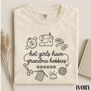 May include: Cream-coloured t-shirt featuring a black graphic of sewing and knitting tools, such as a sewing machine and scissors. The text "hot girls have grandma hobbies" is written in a script font. The shirt is neatly folded.