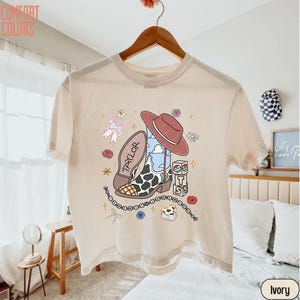 May include: An ivory short-sleeve t-shirt featuring a graphic print of a cowboy boot with a cow print pattern, a red cowboy hat, a blue sky with clouds, and stacked blocks with the letters T, S, and 13. The design includes small flowers, a bow, and a beaded string.