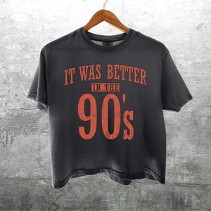 May include: A charcoal grey short-sleeve t-shirt featuring the orange text "IT WAS BETTER IN THE 90'S" printed on the front. The shirt has a crew neckline and a standard fit, displayed on a wooden hanger against a textured grey background.
