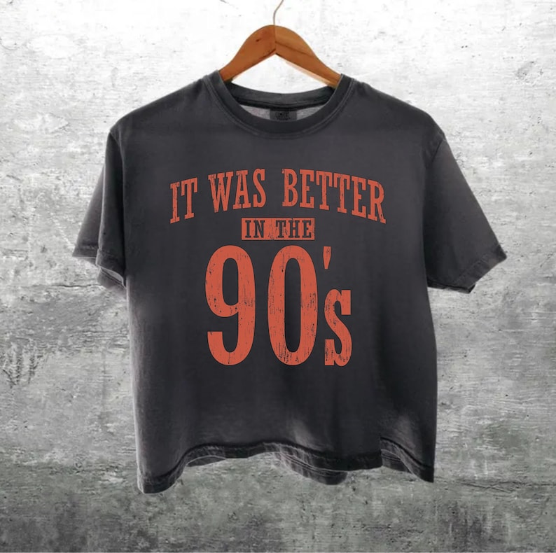 May include: A charcoal grey short-sleeve t-shirt featuring the orange text "IT WAS BETTER IN THE 90'S" printed on the front. The shirt has a crew neckline and a standard fit, displayed on a wooden hanger against a textured grey background.