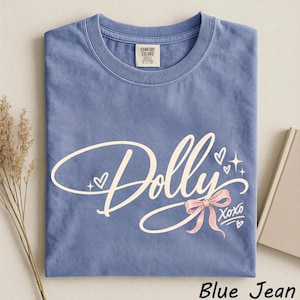 May include: A blue jean colored short-sleeve t-shirt featuring the word Dolly in a cream-colored script font. The design includes a pink bow, small hearts, and star accents, with the text XOXO printed below the bow. A Comfort Colors brand label is visible.