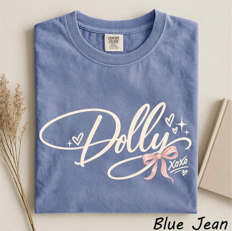 May include: A blue jean colored short-sleeve t-shirt featuring the word Dolly in a cream-colored script font. The design includes a pink bow, small hearts, and star accents, with the text XOXO printed below the bow. A Comfort Colors brand label is visible.