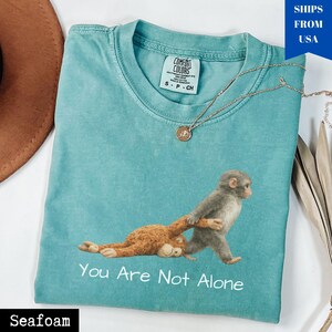 Puede incluir: Camiseta verde mar con un gr&aacute;fico de dos monos y el texto "You Are Not Alone". La camiseta tiene cuello redondo y mangas cortas. Un collar dorado con un colgante est&aacute; sobre la camiseta.