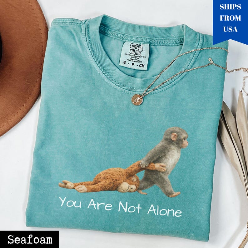 Puede incluir: Camiseta verde mar con un gr&aacute;fico de dos monos y el texto "You Are Not Alone". La camiseta tiene cuello redondo y mangas cortas. Un collar dorado con un colgante est&aacute; sobre la camiseta.