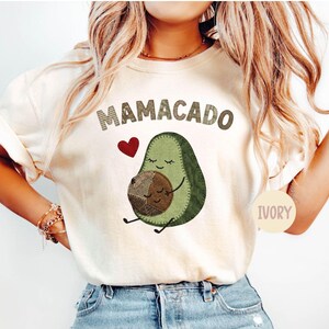 May include: Ivory-colored t-shirt with the word "Mamacado" above a cartoon avocado hugging a baby avocado. A red heart is to the left of the avocado. The word "Ivory" is printed on a circle on the bottom right.