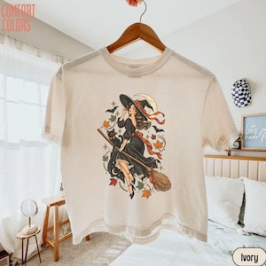 May include: An ivory t-shirt featuring a graphic illustration of a person in a black dress riding a broomstick with a black cat, surrounded by autumn leaves, bats, and a crescent moon. The text COMFORT COLORS is printed in the top left corner.