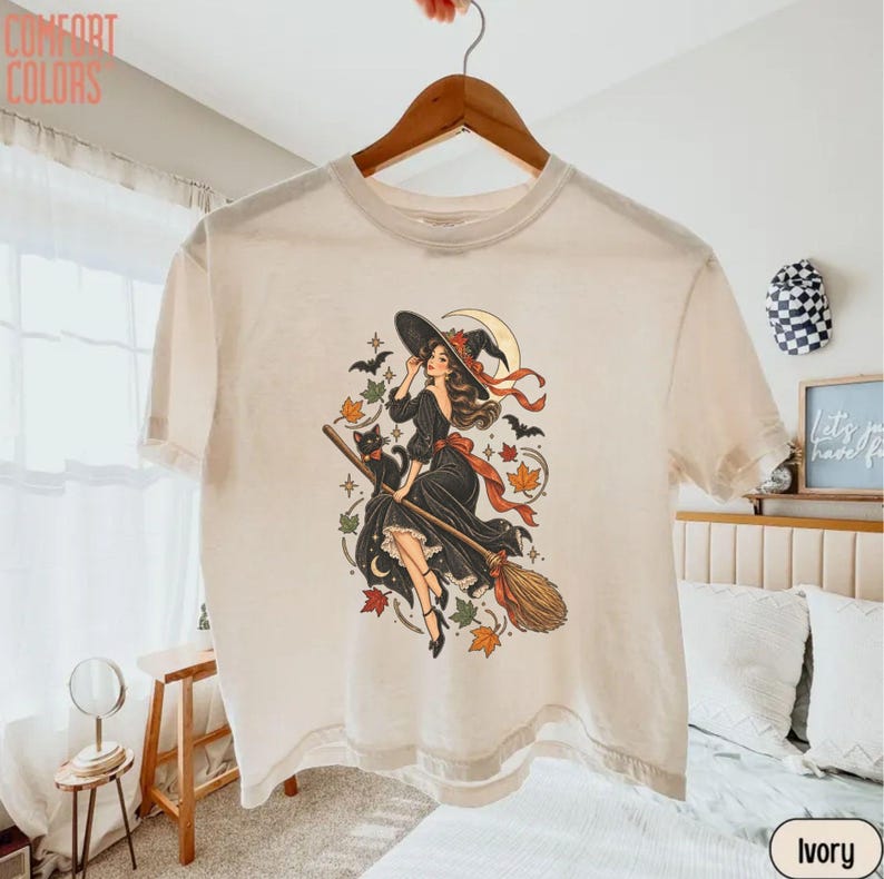 May include: An ivory t-shirt featuring a graphic illustration of a person in a black dress riding a broomstick with a black cat, surrounded by autumn leaves, bats, and a crescent moon. The text COMFORT COLORS is printed in the top left corner.