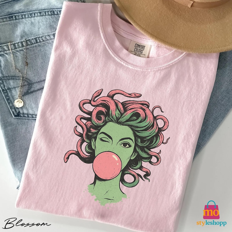 May include: A light pink t-shirt with a graphic of Medusa winking and blowing a bubblegum bubble. The Medusa illustration has green skin and pink hair. The shirt is a Comfort Colors brand.