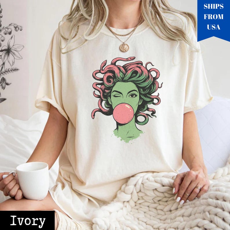 May include: Ivory t-shirt with a graphic of a winking woman with green skin, pink hair, and a bubblegum bubble. The design is centered on the shirt. The shirt is being worn by a person.