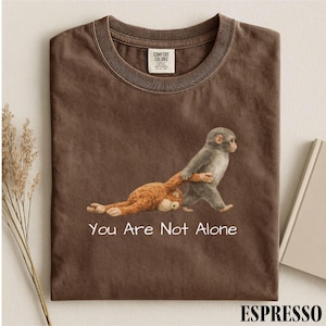 May include: Espresso-colored t-shirt featuring a graphic of two monkeys. One gray monkey is pulling a brown monkey. The text "You Are Not Alone" is printed below the graphic. The shirt is made by Comfort Colors.