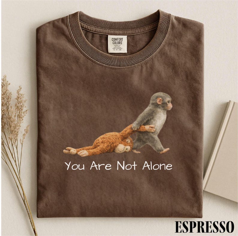 Puede incluir: Camiseta color espresso con un gr&aacute;fico de dos monos. Un mono gris tira de un mono marr&oacute;n. El texto "You Are Not Alone" est&aacute; impreso debajo. La camiseta es de Comfort Colors.