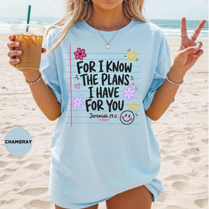 May include: A light blue t-shirt featuring a graphic design that mimics notebook paper with the black text "FOR I KNOW THE PLANS I HAVE FOR YOU" and "Jeremiah 29:11". The design includes colorful pink and purple flowers, yellow stars, and a smiley face.