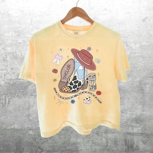 May include: A pale yellow short-sleeve t-shirt featuring a graphic print of a brown cowboy hat, a cow-print boot with the name Taylor, the number 13, and stacked blocks with the letters T, S, and S, surrounded by small flowers, bows, and stars.