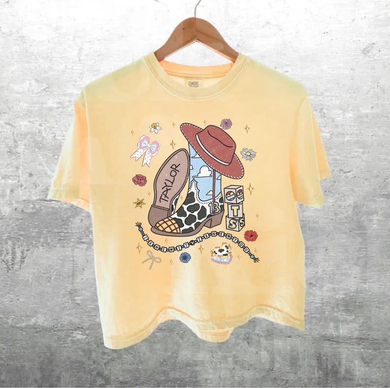 May include: A pale yellow short-sleeve t-shirt featuring a graphic print of a brown cowboy hat, a cow-print boot with the name Taylor, the number 13, and stacked blocks with the letters T, S, and S, surrounded by small flowers, bows, and stars.