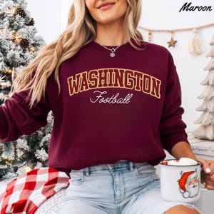 May include: Maroon sweatshirt with "WASHINGTON Football" in gold and white lettering. The sweatshirt is a solid maroon color. A white mug with a Santa Claus graphic is held in the hand.