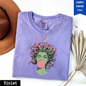 May include: A violet-coloured t-shirt featuring a graphic of Medusa winking and blowing a bubblegum bubble. The Medusa illustration has green skin, pink and green hair, and a pink bubble. A gold necklace is draped across the shirt.