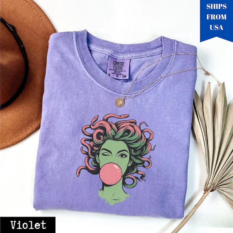 May include: A violet-coloured t-shirt featuring a graphic of Medusa winking and blowing a bubblegum bubble. The Medusa illustration has green skin, pink and green hair, and a pink bubble. A gold necklace is draped across the shirt.