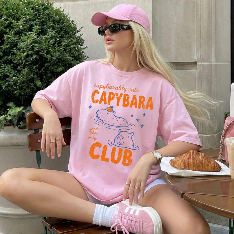 May include: A light pink t-shirt featuring a graphic of two capybaras in water with an orange on one head. The text reads capybarably cute CAPYBARA good vibes only CLUB. The shirt is worn with a matching pink baseball cap and pink sneakers.