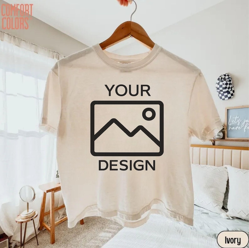 May include: An ivory short-sleeve t-shirt hanging on a wooden hanger. The front of the shirt features the black text 'YOUR' above a black rectangular icon depicting a mountain and sun, with the black text 'DESIGN' printed below the icon.