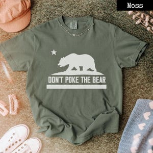 May include: A moss green t-shirt with a white graphic of a bear, a star, and the text "DON'T POKE THE BEAR." The shirt is short-sleeved and has a crew neck. A necklace with small charms is around the neck.