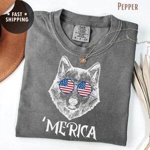Puede incluir: Camiseta gris con una ilustración blanca de un perro con gafas de sol con el diseño de la bandera estadounidense. La palabra "'MERICA" está impresa debajo. Un collar plateado con un colgante está sobre la camisa.