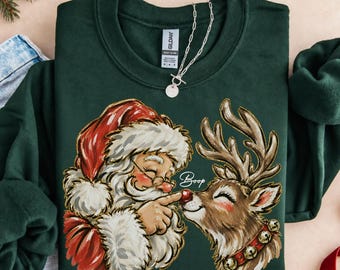 May include: A dark green crewneck sweatshirt featuring a graphic illustration of Santa Claus touching the red nose of a reindeer. The text Boop is written in white script above the reindeer. The garment has a Gildan brand label at the neck.
