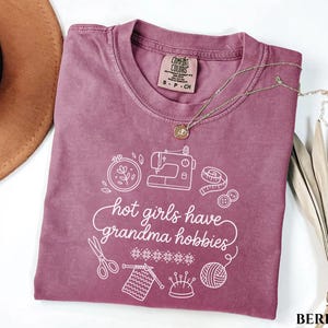 May include: A berry-coloured t-shirt featuring the white text "hot girls have grandma hobbies". The design includes illustrations of sewing tools, knitting needles, and a ball of yarn. A gold necklace and a brown hat are also visible.