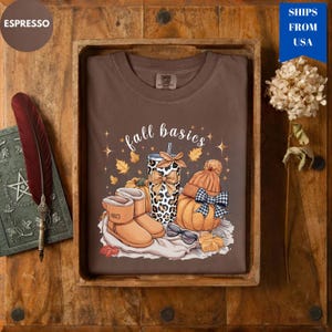 May include: A brown t-shirt with the words "fall basics" and fall-themed graphics, including a pumpkin, boots, and a leopard print tumbler. The shirt is displayed in a wooden tray with a feather and a book.