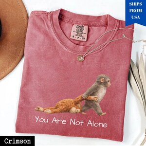 Puede incluir: Camiseta de color carmes&iacute; con un gr&aacute;fico de dos monos, uno caminando y tirando del otro. El texto "You Are Not Alone" est&aacute; impreso debajo del gr&aacute;fico. Un collar plateado con un colgante est&aacute; sobre la camiseta.
