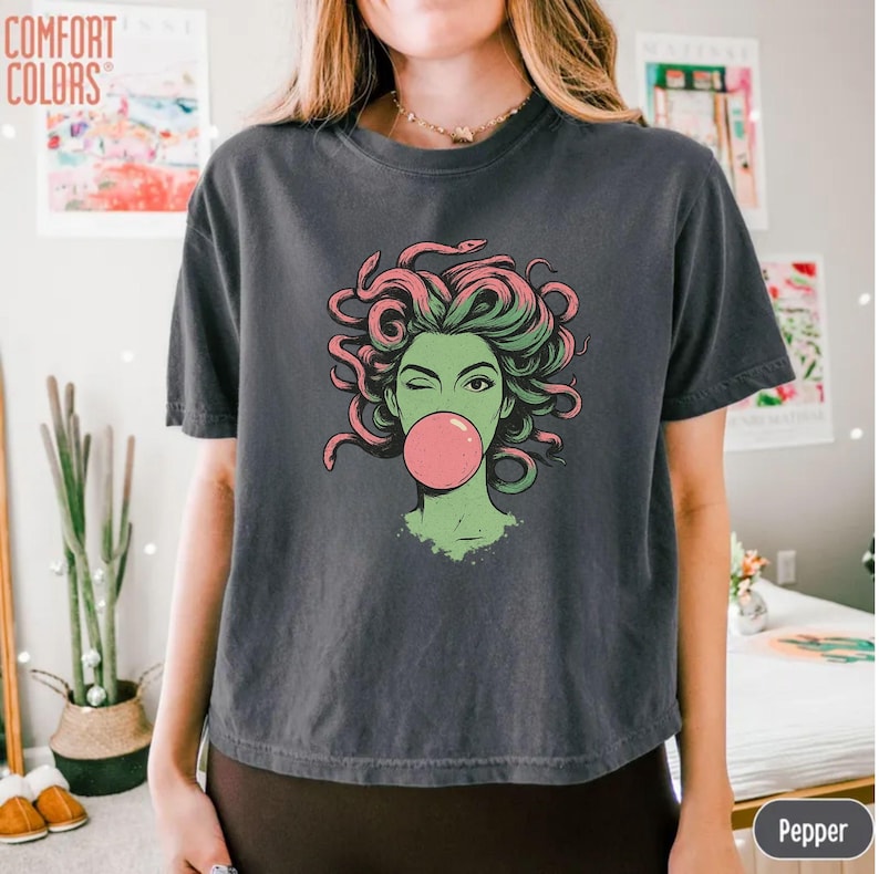 May include: Dark grey t-shirt featuring a graphic of a green-skinned woman with pink and green hair blowing a pink bubble. The graphic is a stylized illustration of Medusa. The words "COMFORT COLORS" are printed in the upper left corner.