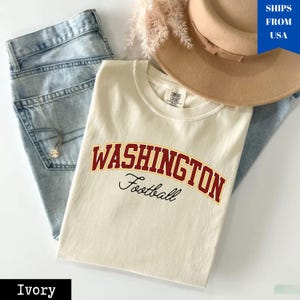 May include: Ivory-colored t-shirt with the word "WASHINGTON" in maroon and gold lettering, and the word "Football" in black script. The shirt is laid on top of light blue jeans, a tan hat, and a necklace.