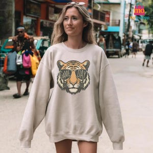 May include: A beige sweatshirt featuring a tiger face design wearing sunglasses with the text "GAME DAY". The tiger has orange, black, and white details. The sweatshirt is a casual style.
