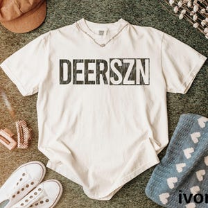 May include: An ivory short-sleeve t-shirt featuring the text DEERSZN in a dark, camouflage-patterned font across the chest. The shirt has a crew neckline and a relaxed fit, presented as a solid-colored garment with a distinct graphic print.
