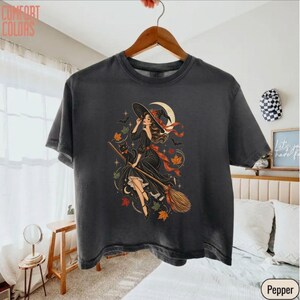May include: A charcoal gray t-shirt featuring a graphic illustration of a witch in a black dress and pointed hat riding a broomstick with a black cat, surrounded by orange and yellow autumn leaves, small stars, and a crescent moon.