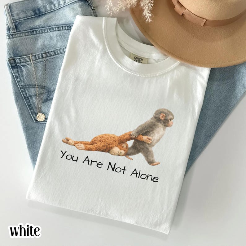 Puede incluir: Camiseta blanca con un gr&aacute;fico de dos monos, uno gris y otro marr&oacute;n, con el texto "You Are Not Alone". La camiseta se muestra sobre unos vaqueros azules con un collar dorado y un sombrero beige.