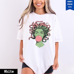 May include: White t-shirt with a graphic of Medusa blowing a bubblegum bubble. The Medusa illustration has green skin, pink and green hair, and is winking. The text "White" is at the bottom of the shirt.