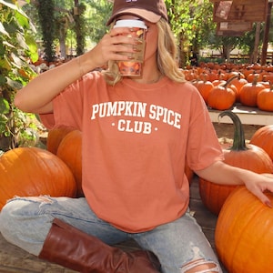 May include: A burnt orange short-sleeve t-shirt featuring the white text PUMPKIN SPICE CLUB printed on the chest. The shirt is worn with light blue distressed denim jeans and brown leather boots, surrounded by numerous orange pumpkins.