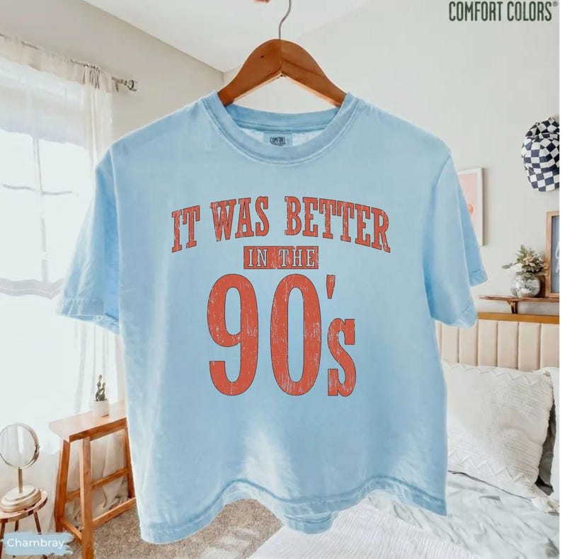 May include: A light blue short-sleeve t-shirt with a crew neckline. The front features distressed red text that reads IT WAS BETTER IN THE 90'S. The shirt is made by Comfort Colors and is displayed on a wooden hanger against a neutral bedroom background.